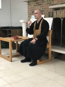 Man in Buddhist monk robes sitting in a chair and gesturing as he discusses a Buddhist concept; small table with a red cup on the side