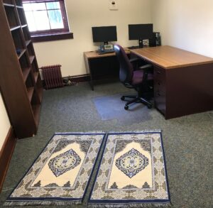 Small office with two prayer rugs, shelves, and desk with rolling chair and computers, facing qibla. 
