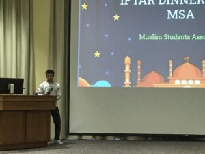 Man holding a microphone while standing in front of a podium and large projector screen with "Iftar Dinner MSA Muslim Students Association" and graphic of minarets.